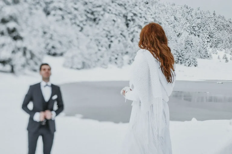 Elena wrapped in a white shawl beside a partly frozen lake, with Vassilis blurred behind her in the snowy landscape