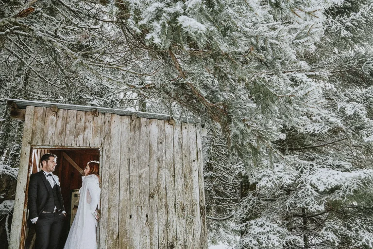 Elena and Vassilis standing in the doorway of a wooden cabin surrounded by snow-covered fir trees