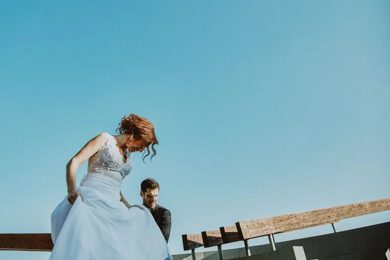Vassilis helping Elena with her wedding dress beneath wooden beams and a clear blue sky