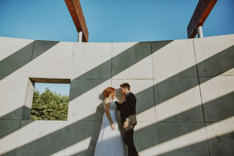 Elena and Vassilis facing each other against a concrete wall crossed by diagonal sunlight and shadows