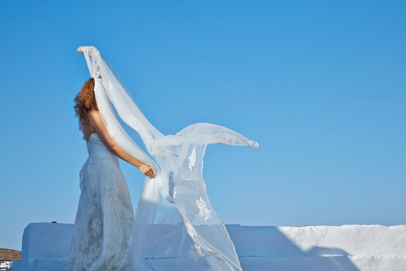 Valmina lifting her lace wedding veil into the breeze against a clear blue sky at Chrisopigi in Sifnos