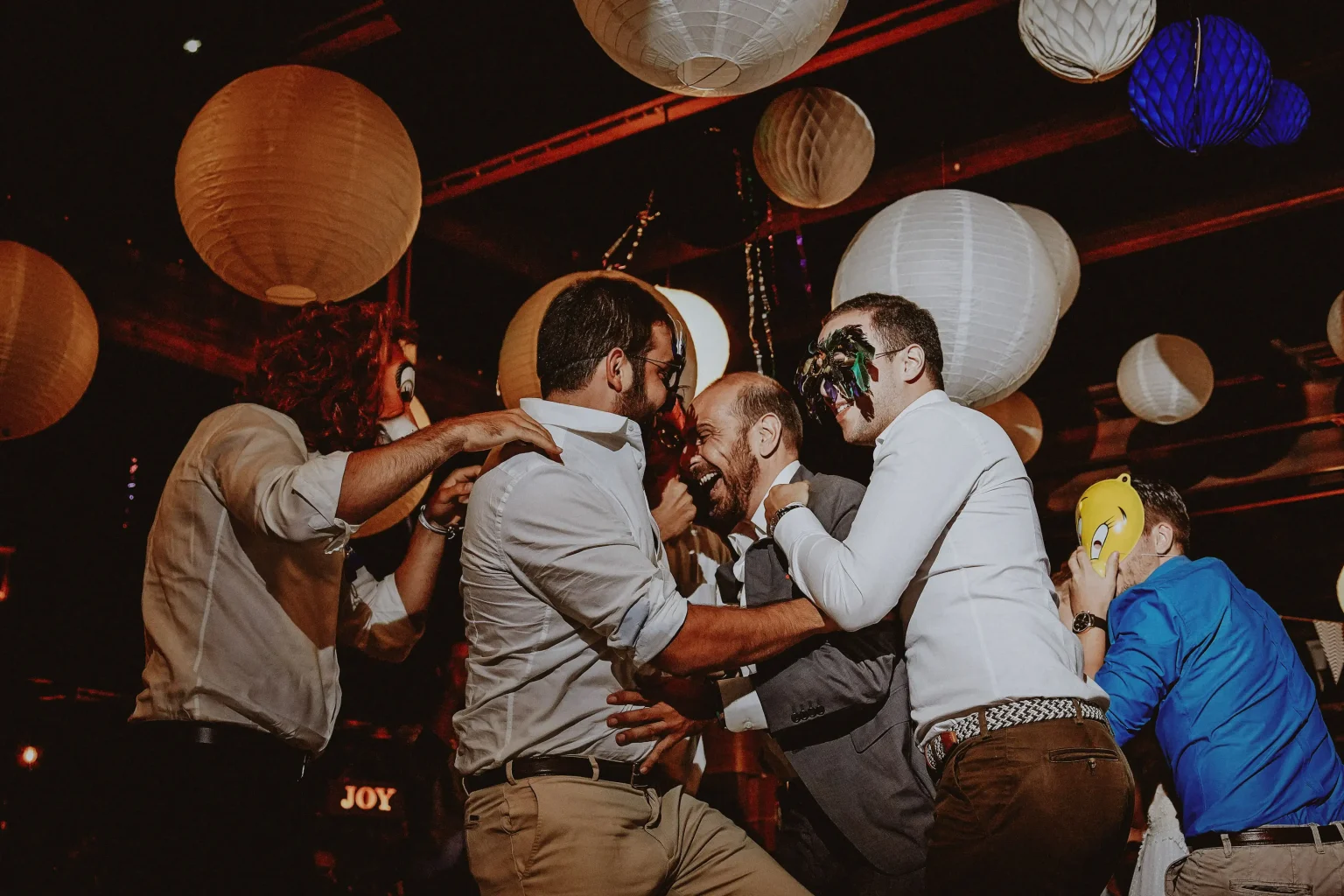 Fanis laughing with friends wearing colourful masks and wigs at the Lazarou Beach wedding reception