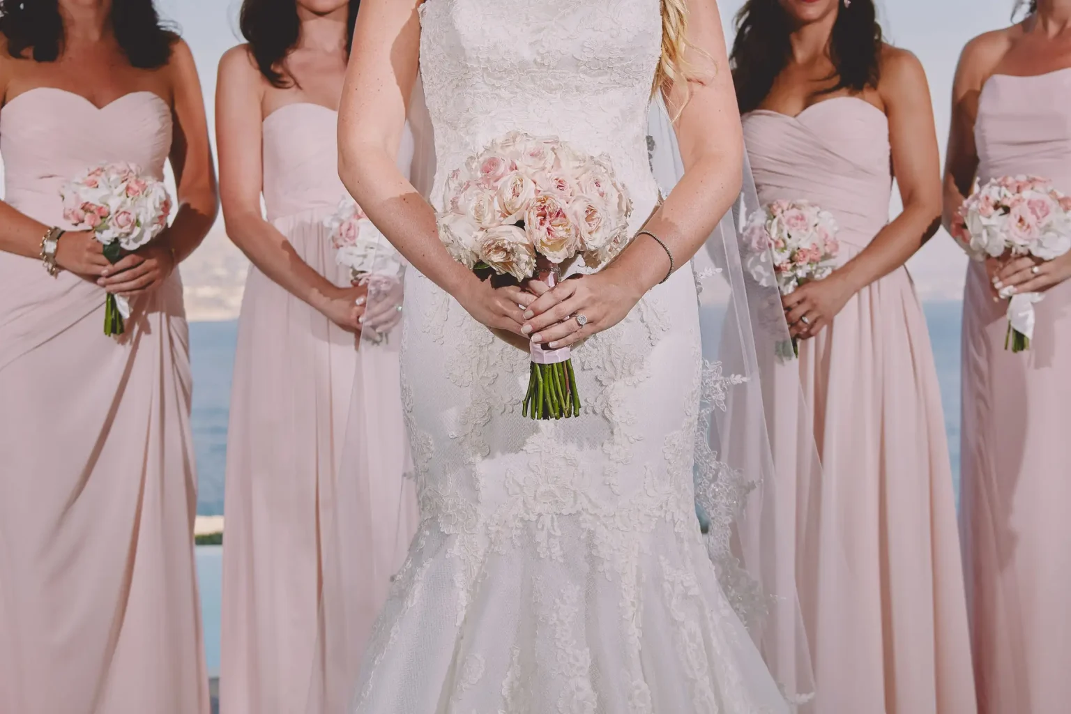 Detail of Abby’s lace wedding gown and rose bouquet, surrounded by bridesmaids in blush dresses
