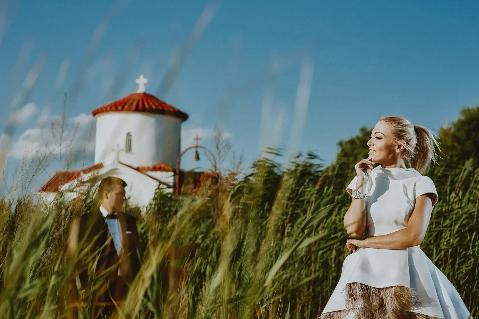 Editorial day-after couple’s portrait of Niki among tall reeds, with Christodoulos with Agios Evplos in the background