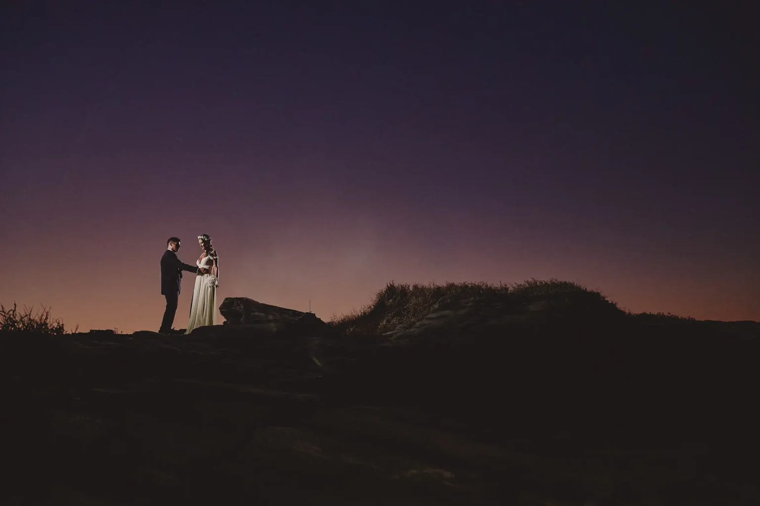Bride and groom Vassiliki and Terry holding hands on the rocks at Chrisopigi beneath a purple twilight sky