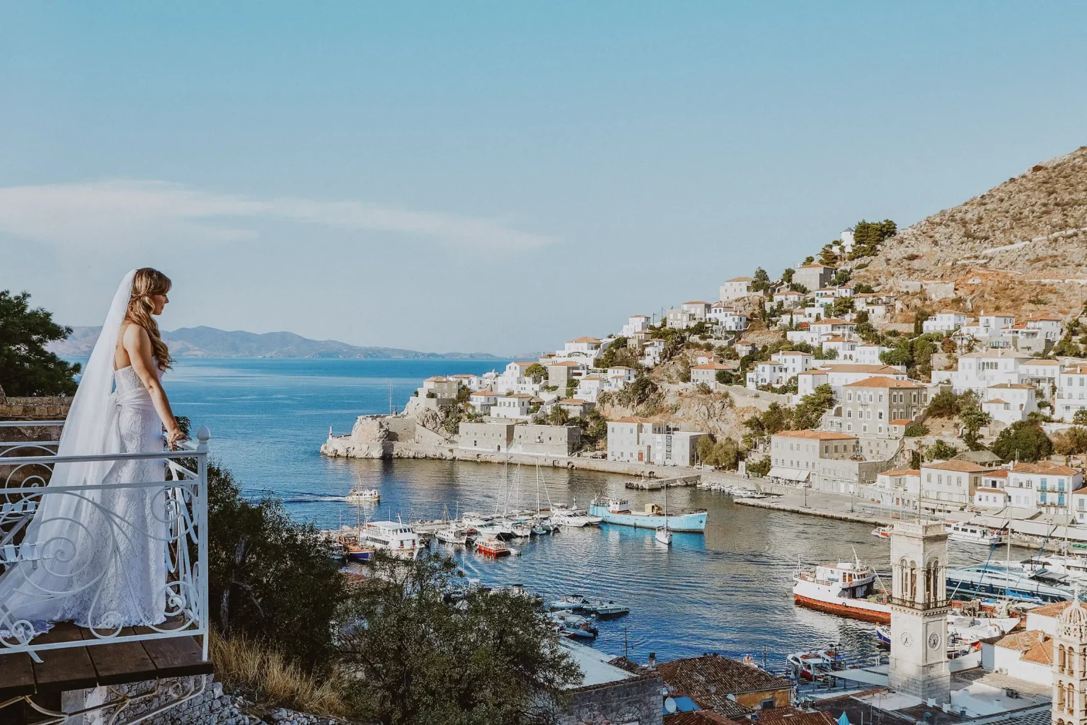 Natalie standing on a balcony in her wedding dress, overlooking Hydra harbour and hillside houses