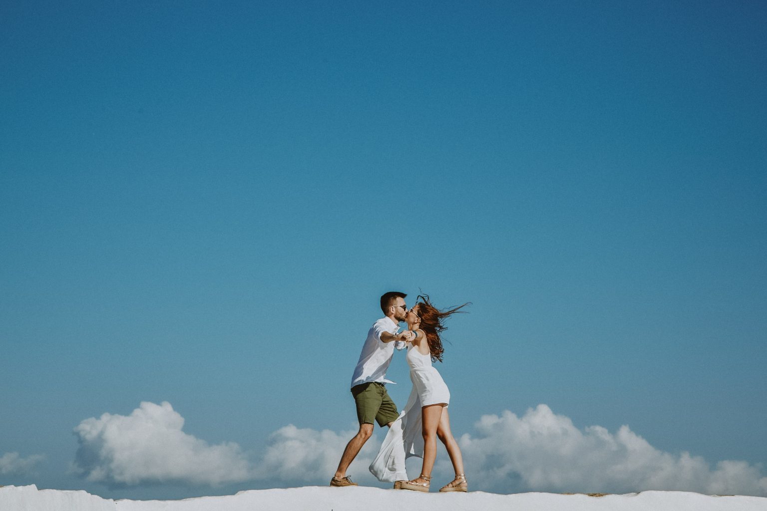 Eliza and Andreas kissing on a whitewashed wall at Chrisopigi during their day-after session beneath a bright blue Sifnos sky