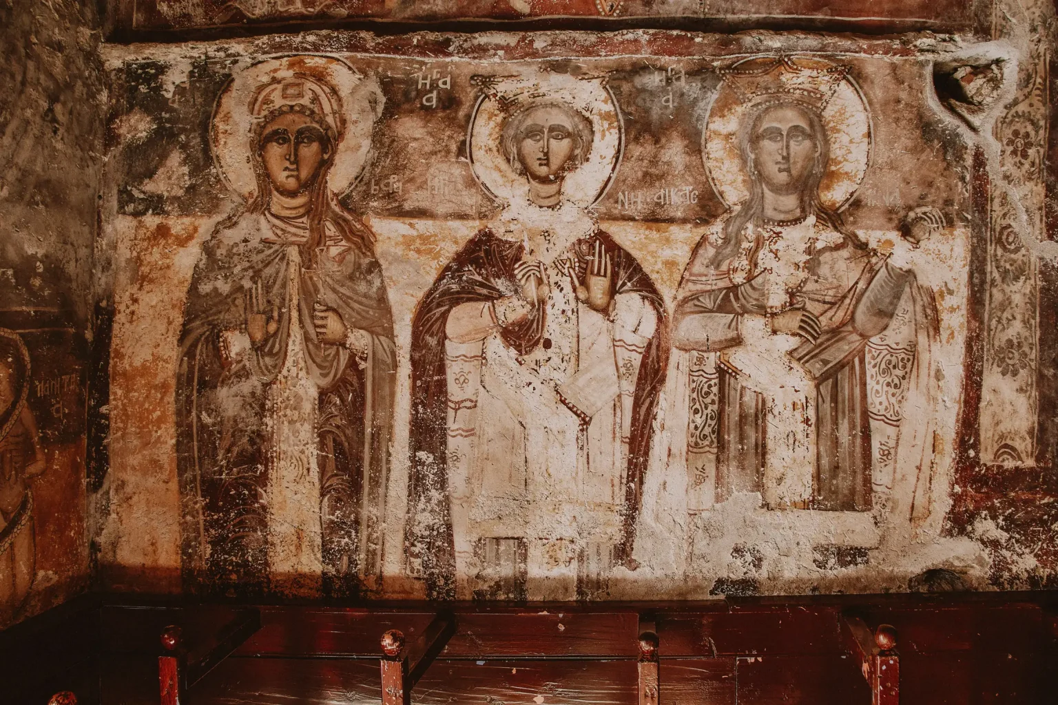 Weathered frescoes of haloed saints above wooden seating inside a church in Ikaria