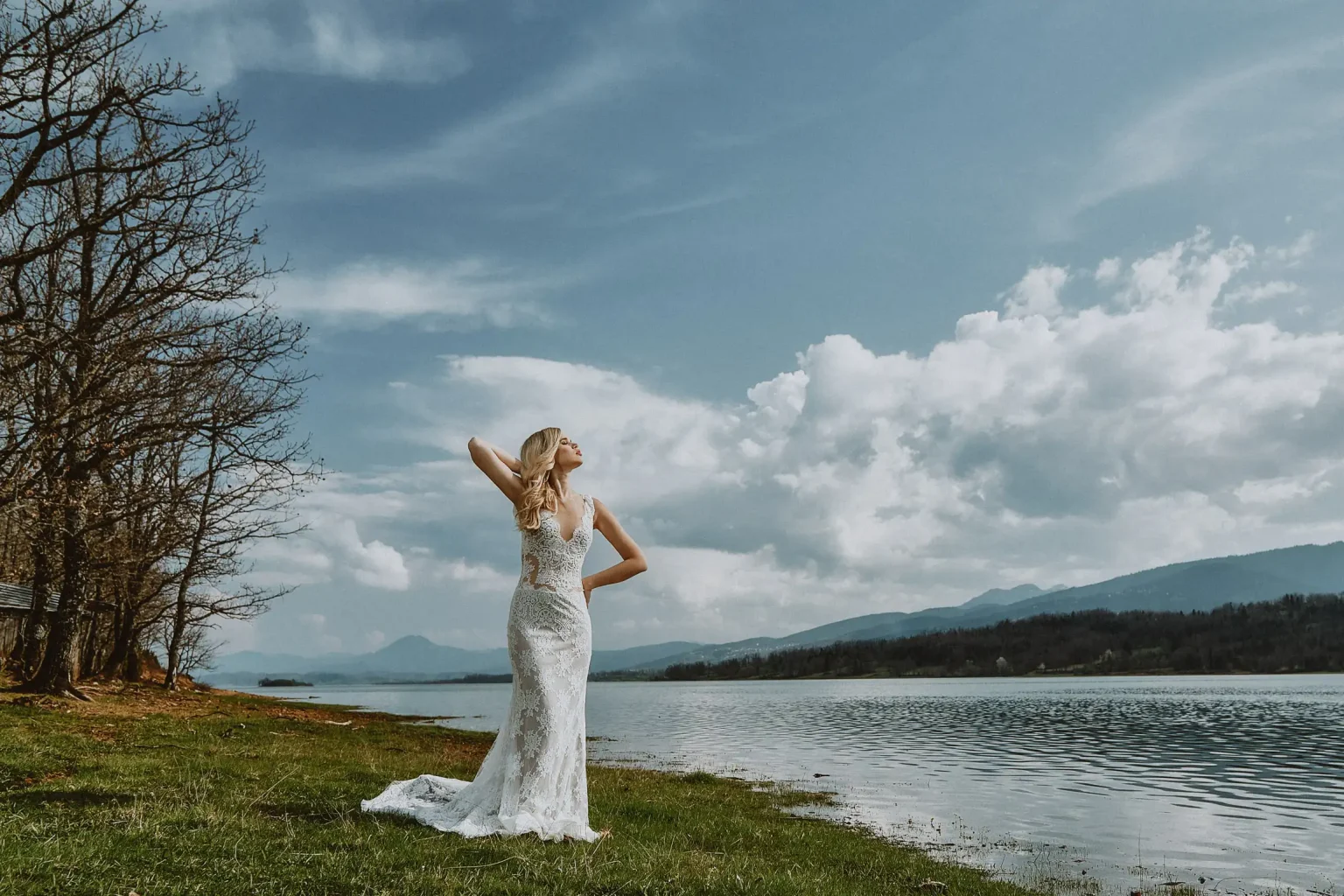 Editorial bridal portrait in a fitted lace wedding dress on the grassy shore of Lake Plastiras beneath a cloudy sky