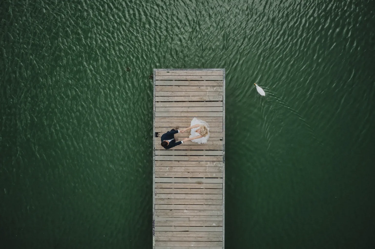 Aerial wedding portrait of Marina and Antonis holding hands on a wooden jetty, with a white bird swimming beside them