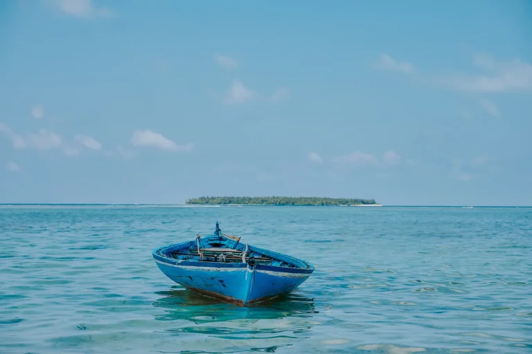 Weathered blue wooden boat floating in a turquoise lagoon, with a palm-covered island on the horizon in the Maldives.