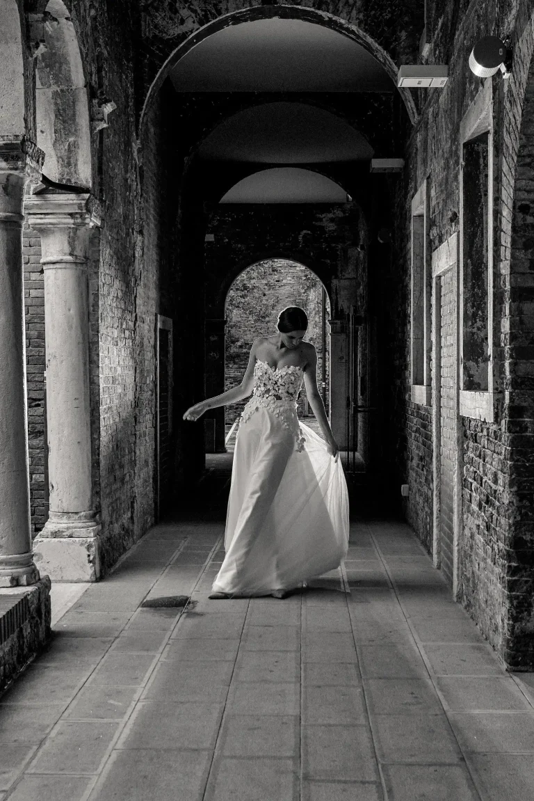 Black-and-white photograph of a bridal model holding the skirt of her gown beneath a series of brick arches in Venice.