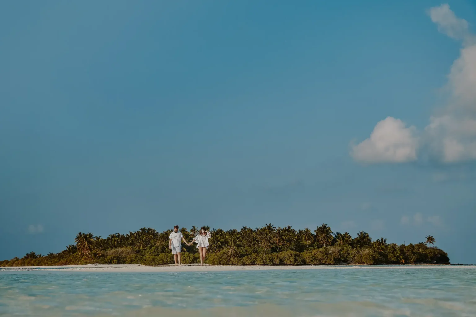 Fay and George holding hands at the water’s edge, with a palm-covered island behind them in the Maldives.