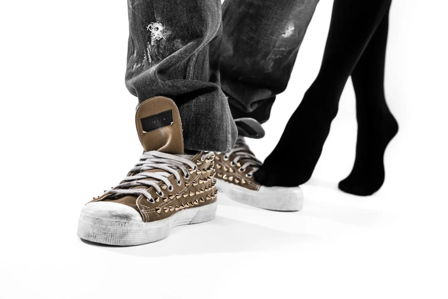 Close-up of Alex's studded sneakers and ripped jeans beside Despoina's black tights during their studio proposal session.